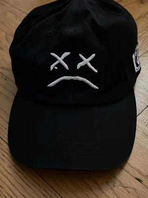 Svart keps med broderad smiley - Säljer en svart keps med böjd skärm och broderad vit smiley med korsade ögon och nedåtvänd mun på framsidan. Kepsen har justerbar rem bak och en patch på sidan. Perfekt för dig som gillar streetwear och vill sticka ut.