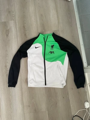 Liverpool träningsjacka Nike grön/vit - Snygg träningsjacka från Nike med Liverpool FC-logga och AXA-sponsor. Jackan är vit med svarta ärmar och detaljer samt en grön panel framtill. Hel dragkedja och två fickor. Perfekt för fotbollsträning eller som sportig streetwear. Den är oanvänd 