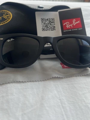 Ray-Ban Wayfarer - Klassiska svarta Wayfarer solglasögon från Ray-Ban med mattsvart båge och mörka glas. Ikonisk design med Ray-Ban-logga på linsen och insidan av skalmen. Kommer med originalfodral och putsduk. Perfekt accessoar för soliga dagar.