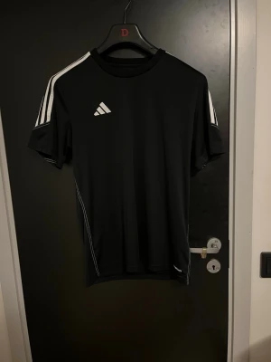 Svart Adidas sporttröja - Snygg svart sporttröja från Adidas med vita ränder på axlarna och logga på bröstet. Tillverkad i ett lätt och snabbtorkande material, perfekt för träning eller gym. Klassisk rund hals och korta ärmar för maximal rörelsefrihet.