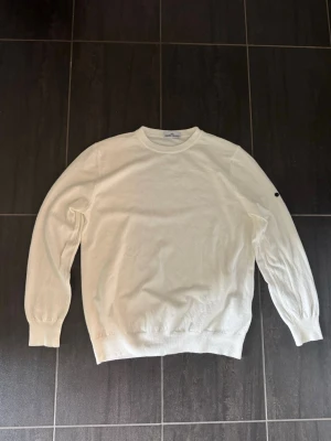 Vit Stone Island merinotröja - Vit sweatshirt från Stone Island med klassisk rund hals och ribbade muddar. Tröjan har den ikoniska Stone Island-loggan på vänster ärm. Perfekt för en clean och stilren look.