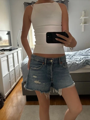 Blå denimshorts med fransig kant - Säljer ett par blå denimshorts med fransig kant och slitna detaljer framtill. Klassisk femficksmodell med knappgylf och avslappnad passform. Perfekta till sommaren och lätta att matcha med olika toppar. Köpte för 329 säljer för 160