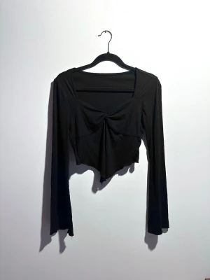 Svart croppad långärmad topp - Trendig svart croppad topp med lång ärm och twistad detalj framtill. Toppen har en asymmetrisk nederkant och är tillverkad i mjukt stretchigt material som sitter snyggt på kroppen. Perfekt för dig som gillar en edgy och modern stil.