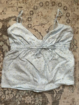 Ljusblå blommig linnetopp med knyt - Supersöt ljusblå linnetopp med tunna axelband och små vita blommor. Toppen har en söt knytning i midjan och v-ringad hals. Perfekt för varma dagar och ger en romantisk vibe till din outfit. Onesize men passar xs/s