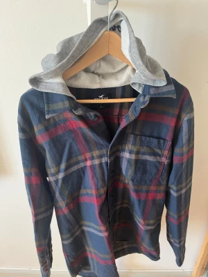 Hollister Overshirt  - Sjukt snygg vintage overshirt från Hollister med rutig skjortaktig tröja med en grå huva med texten ”Hollister” på. Mycket bra skick, storlek L. Skriv vid eventuella funderingar😁 fraktar inom 1-2 dagar 