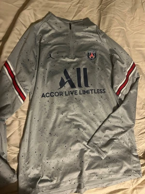 PSG x Jordan grå träningströja - Snygg grå långärmad träningströja från PSG x Jordan med svartprickigt mönster, halv dragkedja och klubbmärke på bröstet. Röda och blå detaljer på ärmarna och trycket 'Accor Live Limitless' framtill. Perfekt för fotbollsträning eller chill.