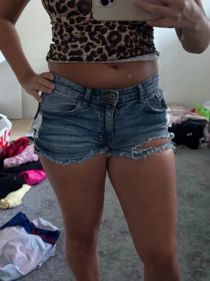 Blå jeansshorts med fransar - Säljer ett par blå jeansshorts med råa, fransiga kanter och slitningar. Klassisk femficksmodell med knapp och dragkedja framtill. Perfekta för varma dagar och ger en avslappnad vibe.