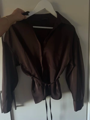 Mörkbrun blus med knytband zara - Snygg mörkbrun blus med långa ärmar och knytband i midjan. Blusen har klassisk krage och en lätt glansig finish som ger en elegant touch. Perfekt att styla med jeans eller kostymbyxor för en trendig look. Använd en gång, från zara