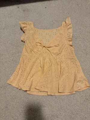 Gul omlottopp med volangärmar - Söt gul omlottopp med broderade hål och volangärmar. Toppen har v-ringning och knyt framtill, vilket ger en feminin och somrig känsla. Perfekt till jeans eller kjol för en fräsch look.