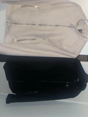 2 för 1 Beige och svart äkta mockajackor - Snygg beige och svart mockajacka med raka linjer och silverfärgade dragkedjor framtill och vid fickorna. Jackan har lång ärm och en stilren, minimalistisk look. Perfekt för dig som gillar en clean och modern stil.