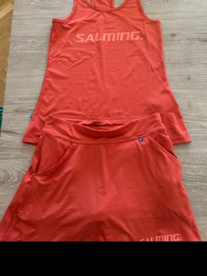 Orange padelset  Salming - Snyggt orange padel/tennis set från Salming. Setet har ett sportigt linne med Salming-logga på bröstet och en kjol med fickor och logga nertill. Perfekt för träning eller padel. Materialet är mjukt och stretchigt för maximal rörelsefrihet.