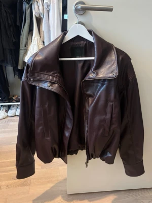 Vinröd skinnjacka med bred krage - Snygg mörkbrun skinnjacka från Alohas med bred krage och dragkedja framtill. Jackan har resår i midjan och vid ärmsluten för en cool, avslappnad passform. Perfekt för dig som vill ha en trendig och tidlös look.