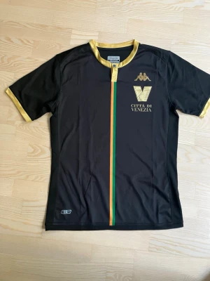 Svart Venezia fotbollströja Kappa - Snygg svart fotbollströja från Kappa med gulddetaljer och tryck 'Città di Venezia' på bröstet. Tröjan har korta ärmar, guldfärgade muddar och en rand i grönt, orange och rött längs mitten.