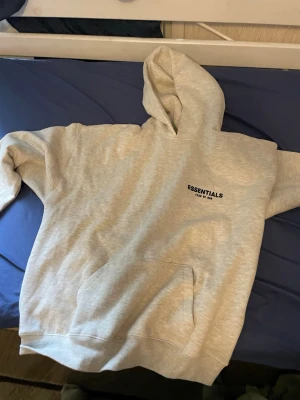 Grå Essentials Fear of God hoodie - Säljer en ljusgrå hoodie från Essentials Fear of God med svart logga på bröstet och stor text på ryggen. Hoodien har en klassisk känguruficka och huva. Tillverkad i mjukt bomullsmaterial som är perfekt för en avslappnad streetwear-look.