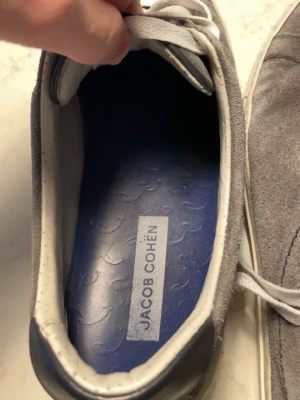 Gråa sneakers från Jacob Cohën - Snygga sneakers från Jacob Cohën i grå mocka med vita snören och blå innersula. Skorna har en klassisk låg siluett och vita detaljer runt sulan. Perfekta för dig som gillar stilrena och tidlösa sneakers. Säljer dessa eftersom jag har använt dom ett tag och är ute efter något nytt