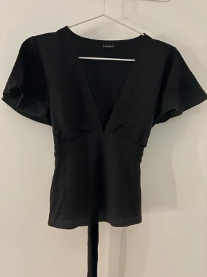 Svart topp med djup v-ringning Gina Tricot - Svart topp från Gina Tricot med djup v-ringning och korta, vida ärmar. Figurnära passform med markerad midja och knytband bak. Perfekt för en trendig och stilren look.