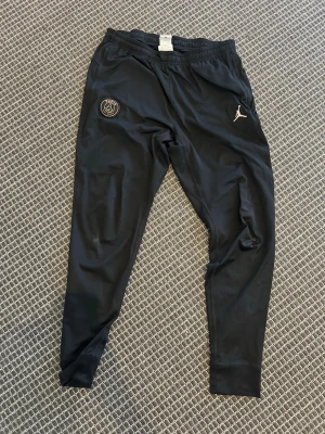 Svarta PSG x Jordan träningsbyxor - Säljer ett par svarta träningsbyxor från Jordan i samarbete med Paris Saint-Germain. Byxorna har PSG-logga i guld på ena benet och Jumpman-logga på andra. Tillverkade i ett mjukt, stretchigt material med smal passform och resår i midjan.