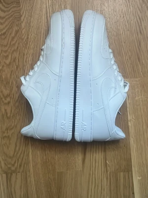 Nike Air Force 1 vita - Klassiska helvita Nike Air Force 1 sneakers i lågt utförande. Skorna har en clean design med perforerad tå, vita skosnören och den ikoniska swoosh-loggan på sidan. Tillverkade i slätt läder med en rejäl sula för skön komfort.