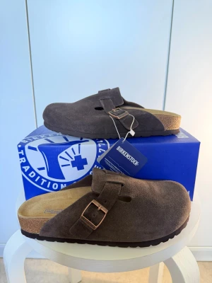 Birkenstock Boston Mocha tofflor - Säljer ett par Birkenstock Boston tofflor i färgen mocha. De är gjorda i mjukt mocka med justerbart spänne i metall och har en klassisk korksula för extra komfort. Perfekta för dig som gillar stilrena och bekväma skor.