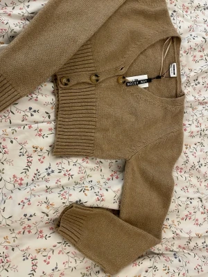 Beige cropped kofta från Noisy May - Snygg beige cropped kofta från Noisy May med ribbade muddar och knappar framtill. Koftan har långa ärmar och är gjord i ett mjukt stickat material som passar perfekt till jeans eller kjol.