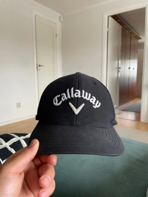 Svart Callaway keps med brodyr - Snygg svart keps från Callaway med vit broderad logga framtill och Odyssey-detalj baktill. Klassisk böjd skärm och justerbar rem bak. Perfekt för golfbanan eller som streetwear-accessoar. Tillverkad i slitstarkt tyg.