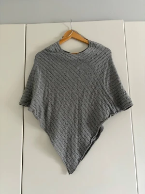 Grå stickad poncho  - Snygg grå stickad poncho med snedskuren nederkant och diagonalt ribbat mönster. Ponchon har en rund halsringning och är perfekt att slänga över en tröja för extra värme. Materialet känns mjukt och följsamt, vilket gör den både bekväm och stilren.