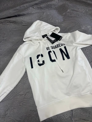 Vit hoodie DSQUARED2 ICON - Snygg vit hoodie från DSQUARED2 med stor svart 'ICON'-text på bröstet och justerbar huva med dragsko. Klassisk passform med ribbade muddar och nederkant. Perfekt för dig som gillar streetwear och vill sticka ut med ett trendigt plagg.
