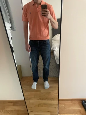 Korallfärgad pikétröja från Ralph Lauren - Säljer en snygg korallfärgad pikétröja från Ralph Lauren med klassisk krage och korta ärmar. Tröjan har den ikoniska blå loggan broderad på bröstet och är tillverkad i mjuk bomull. Perfekt för en avslappnad och stilren look. PS De strå M men är större, som en L