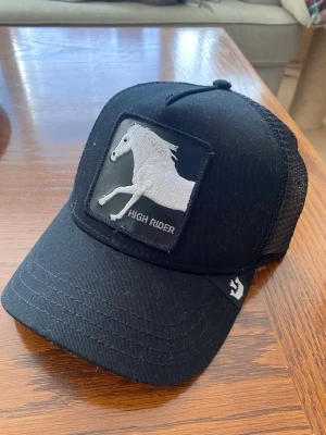 Svart truckerkeps med hästmotiv - Säljer en svart truckerkeps från Goorin Bros med mesh baktill och böjd skärm. Framtill finns ett broderat patch med en vit häst och texten 'HIGH RIDER'. Perfekt för dig som gillar djur och streetwear.