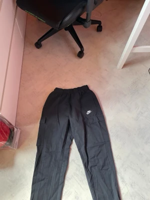 Svarta cargobyxor från Nike - Säljer ett par svarta cargobyxor från Nike med elastisk midja och flera praktiska fickor på sidorna. Byxorna har en avslappnad passform och är perfekta för en streetwear-look. Tillverkade i ett slitstarkt material som känns bekvämt hela dagen.