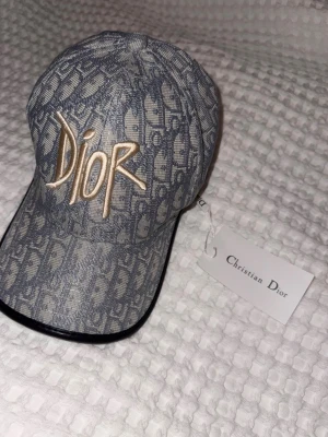 Monogram keps från Dior i denim - Snygg keps från Dior i blått denimtyg med ikoniskt monogrammönster och stor broderad Dior-logga i beige framtill. Böjd skärm med svart kant och justerbar rem bak. Perfekt accessoar för en trendig streetwear-look.
