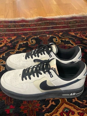 Nike Air Force 1 pris diskuterbar  - Snygga Nike Air Force 1 sneakers i beige mocka med svarta detaljer och svart sula. Klassisk låg modell med svart swoosh på sidan och svarta skosnören. Perforerad tå för extra ventilation och stilren look.