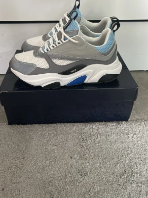 sneakers i vitt, grått och blått - Snygga sneakers med chunky sula i vitt, grått och blått. Ovandelen är i mesh och läder med blå detaljer och svarta inslag. Skorna har vita snören, svart och vit hälflik samt märkning på sidan. Perfekta för dig som gillar exklusiv streetwear.