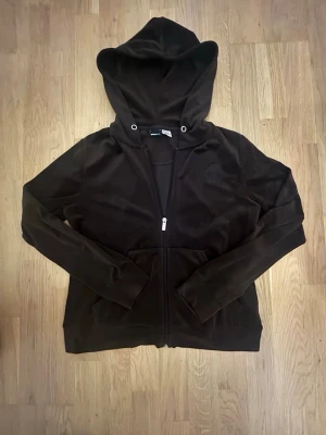 Mörkbrun velourhoodie från Ginatricot - Mjuk och skön mörkbrun hoodie i velour från Ginatricot. Dragkedja framtill, två fickor och stor huva med metallöljetter. Perfekt för en chill och avslappnad stil. Klassisk passform och riktigt cozy material. Snörena är borta