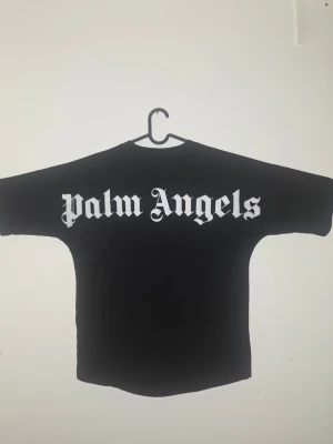 Svart Palm Angels t-shirt - Säljer en svart t-shirt från Palm Angels med stor vit logga i gotisk stil på ryggen och mindre logga i nacken. T-shirten har korta ärmar och en loose passform, perfekt för en streetwear-look.