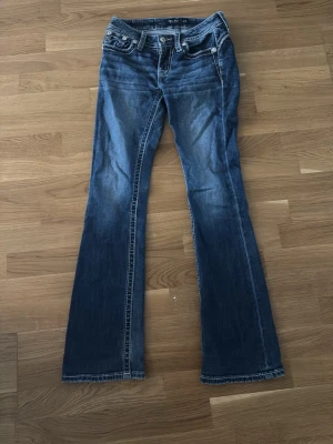 Blå bootcut jeans med broderade fickor - Snygga blå bootcut jeans med slitningar och broderade bakfickor från Miss me. Jeansen har medel hög midja, klassisk femficksdesign och dekorativa detaljer på fickorna. Perfekta för en avslappnad och trendig look. Midja 74  innerbenet längd 79