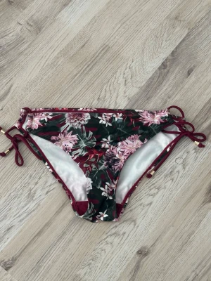 Blommiga bikiniunderdelar med snörning - Snygga bikiniunderdelar med blommigt mönster i vinrött, grönt och rosa. Modellen har snörning i sidorna med guldfärgade detaljer och en bekväm passform. Perfekt för sommaren och strandhäng!