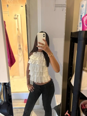 Vit volang topp med knyt i nacken - Supersöt vit topp med flera lager volanger och knyt i nacken. Toppen är ärmlös och har en halterneck-modell som ger en luftig och trendig look. Perfekt för varma dagar och att styla med jeans eller kjol.
