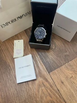 Emporio Armani silverfärgad klocka - Stilren klocka från Emporio Armani med silverfärgat armband i metall och mörkblå urtavla. Klockan har kronograf-funktion, datumvisning och klassisk rund form. Levereras i originalask med manual och garantikort.