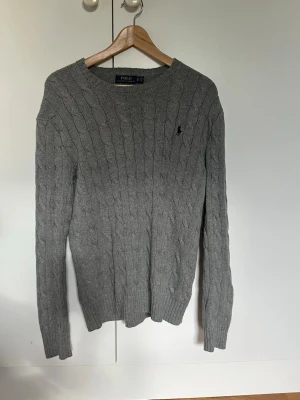 Grå kabelstickad tröja Polo Ralph Lauren - Kabelstickad grå tröja från Polo Ralph Lauren med rund halsringning och diskret broderad logga på bröstet. Långärmad modell i mjukt material som passar perfekt till jeans. Sitter som S/M