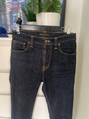 Mörkblå jeans från Nudie Jeans - Säljer ett par mörkblå jeans från Nudie Jeans med klassisk femficksmodell och kontrastsömmar i orange. Jeansen har rak passform och är tillverkade i kraftig denim. Perfekta för dig som gillar stilrena och tidlösa jeans.