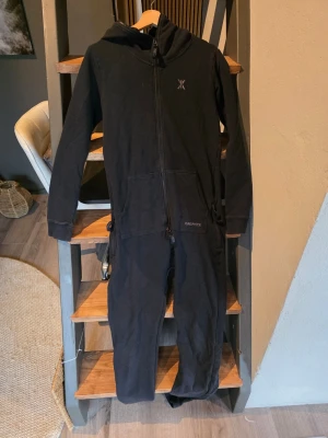 Svart onepiece med huva - Mysig svart onepiece från Onepiece med dragkedja framtill, huva och stora fickor. Perfekt för chill hemma eller när du vill vara extra bekväm. Långärmad och helkroppsdesign med diskret logga på bröstet och ryggen.