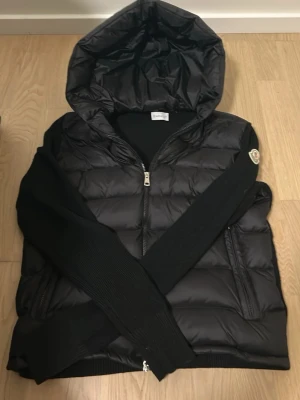 Svart dunjacka med huva från Moncler - Snygg svart dunjacka från Moncler med stickade ärmar och stor huva. Jackan har dragkedja framtill, två fickor och Moncler-logga på ena ärmen. Perfekt för kalla vinterdagar och riktigt trendig look. Pris kan diskuteras vid snabbt affär