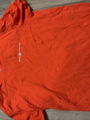 Orange t-shirt från Sail Racing - Snygg orange t-shirt från Sail Racing med vit logga tryckt på bröstet. Klassisk passform och rund halsringning. Tillverkad i mjuk bomull som känns skön mot huden. Perfekt för dig som gillar sportig och enkel stil.