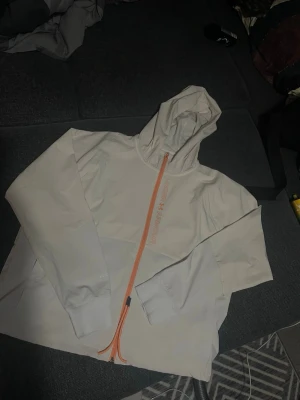  Under Armour - Snygg vit  vind jacka från Under Armour med huva och sneddraget orange blixtlås framtill. Tillverkad i ett mjukt och stretchigt material som känns sportigt och modernt. Perfekt för dig som gillar en clean och stilren look med en twist. Perfekt för friluftsldag eller liknade 