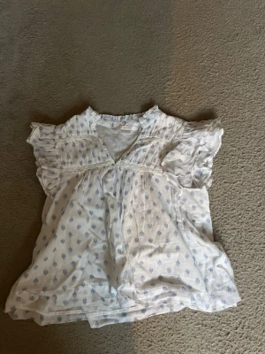 Vit blus med volangärmar och blått mönster - Tryck gärna köp nu🩷🩷 Söt vit blus med volangprydda korta ärmar och diskret blått mönster över hela plagget. Blusen har v-ringning och lätt rynkade detaljer framtill för en luftig och romantisk känsla. Perfekt till jeans eller kjol för en avslappnad men stilsäker look.