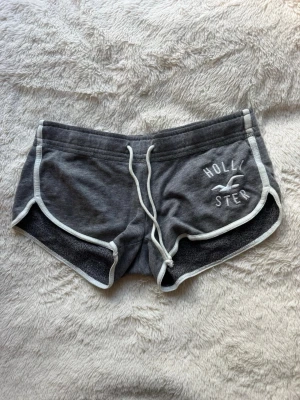 Lågmidjade grå shorts från Hollister med snörning - Lågmidjade grå shorts från Hollister med vit snörning i midjan och vita kanter. Klassisk logga broderad framtill. Perfekta för chill dagar eller träning. Mjukt material som känns skönt mot huden.