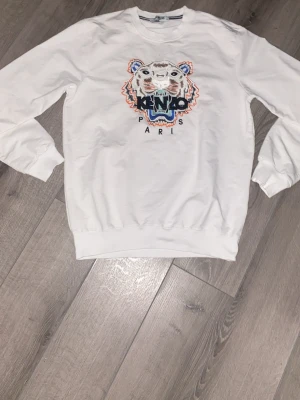 Vit Kenzo sweatshirt med tigertryck - Säljer en vit sweatshirt från Kenzo med det ikoniska broderade tigerhuvudet och texten 'Kenzo Paris' på bröstet. Tröjan har rund hals, långa ärmar och ribbade muddar. Perfekt för dig som gillar streetwear och vill sticka ut med en klassisk design.