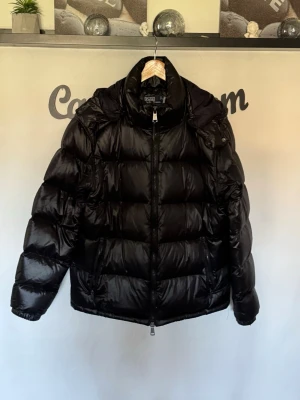Svart dunjacka från Polo Ralph Lauren - Snygg svart dunjacka från Polo Ralph Lauren med glansig finish och stor huva. Jackan har breda kanaler, dragkedja framtill och två sidofickor. Perfekt för kalla vinterdagar och ger en riktigt cool streetwear-look.