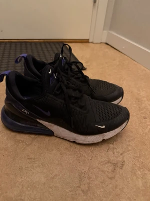 Nike Air Max 270 svarta sneakers - Snygga svarta Nike Air Max 270 sneakers med lila detaljer och vit sula. Skorna har en luftig mesh-överdel, klassisk Nike-logga på sidan och synlig Air-enhet i hälen för extra komfort. Perfekta för dig som vill ha både stil och funktion i vardagen.